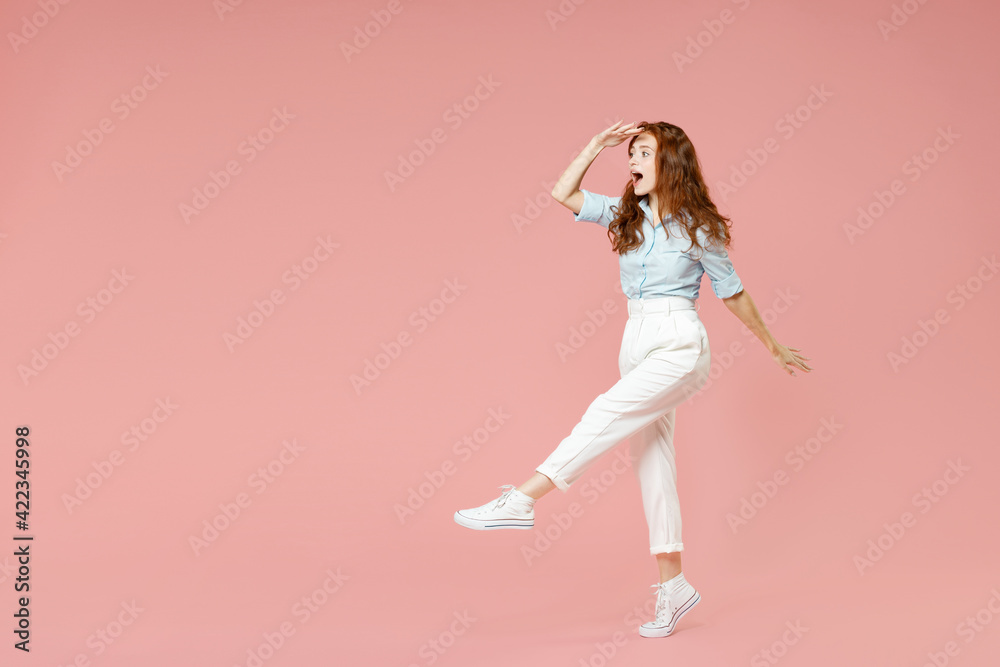 Full length young student optimistic smiling happy redhead woman 20s wear blue shirt pants holding hand at forehead looking far away distance isolated on pastel pink color background studio portrait.