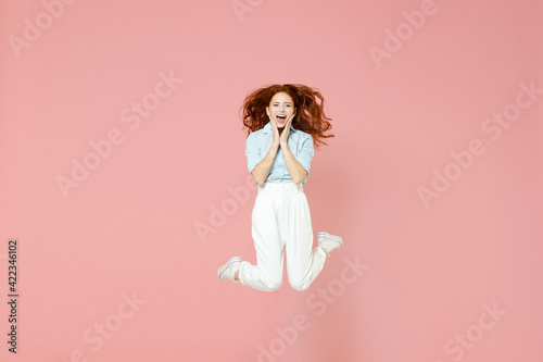 Full length young student amazed surprised impressed shocked excited happy redhead woman 20s wearing blue shirt pants jumping high hold face isolated on pastel pink color background studio portrait