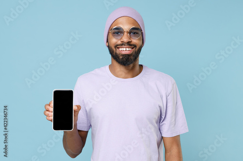 Young smiling happy satisfied black african man in violet t-shirt purprle hat glasses show mobile cell phone, blank screen workspace area mock up isolated on pastel blue background studio portrait photo