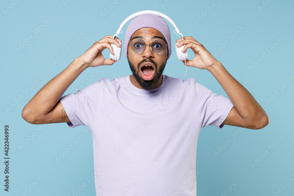 Young surprised shocked impressed unshaven black african man 20s wear violet t-shirt hat glasses listening to music with took off headphones isolated on pastel blue color background studio portrait Young surprised shocked impressed unshaven black african man 20s wear violet t-shirt hat glasses listening to music with took off headphones isolated on pastel blue color background studio portrait