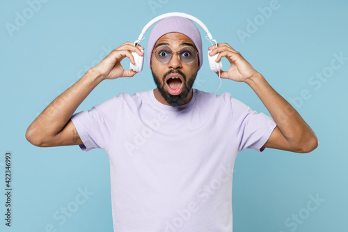 Young surprised shocked impressed unshaven black african man 20s wear violet t-shirt hat glasses listening to music with took off headphones isolated on pastel blue color background studio portrait