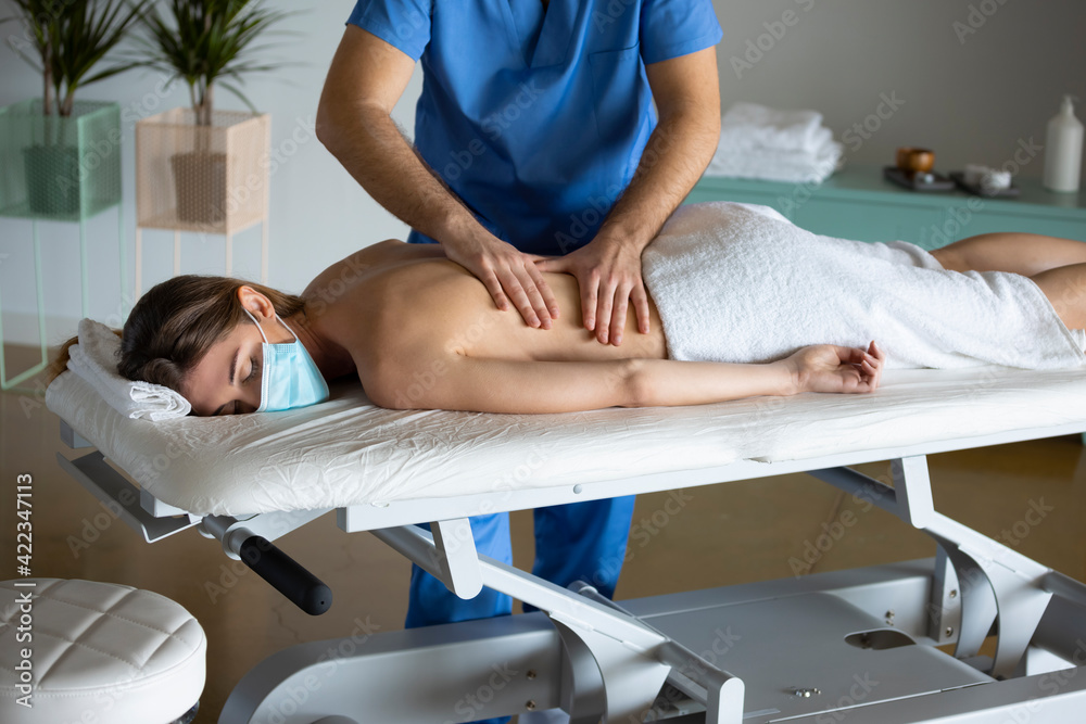 Male physiotherapist giving a massage to a young woman wearing surgical mask and lying down on a stretcher bare back, manual therapy for backache in a health and wellness center.