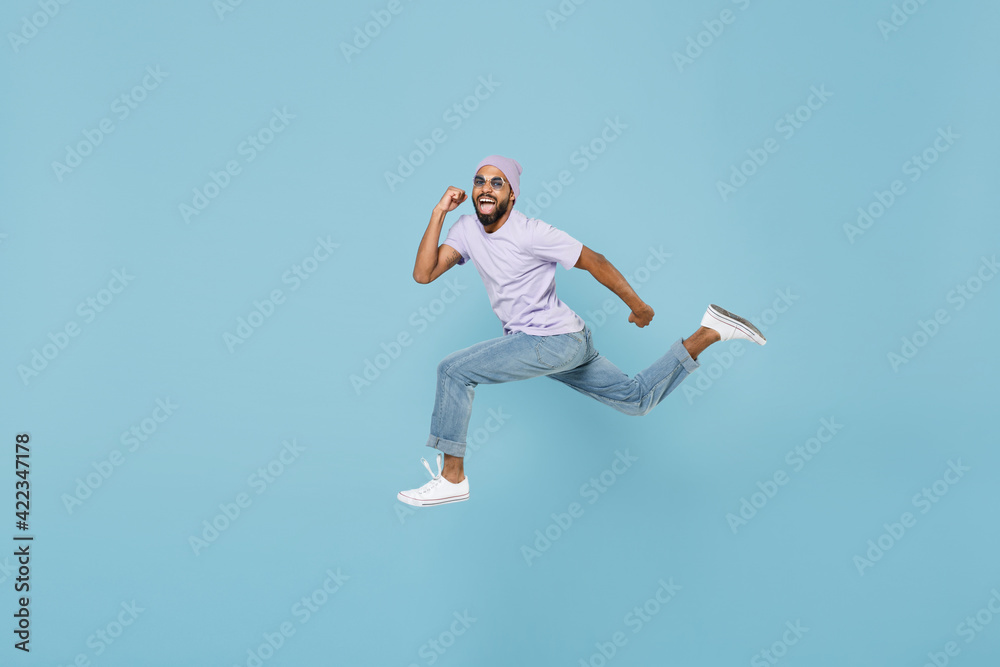 Full length of young smiling positive confident black african man 20s in violet t-shirt hat glasses jump high run fast do winner gesture clench fist isolated on pastel blue background studio portrait.
