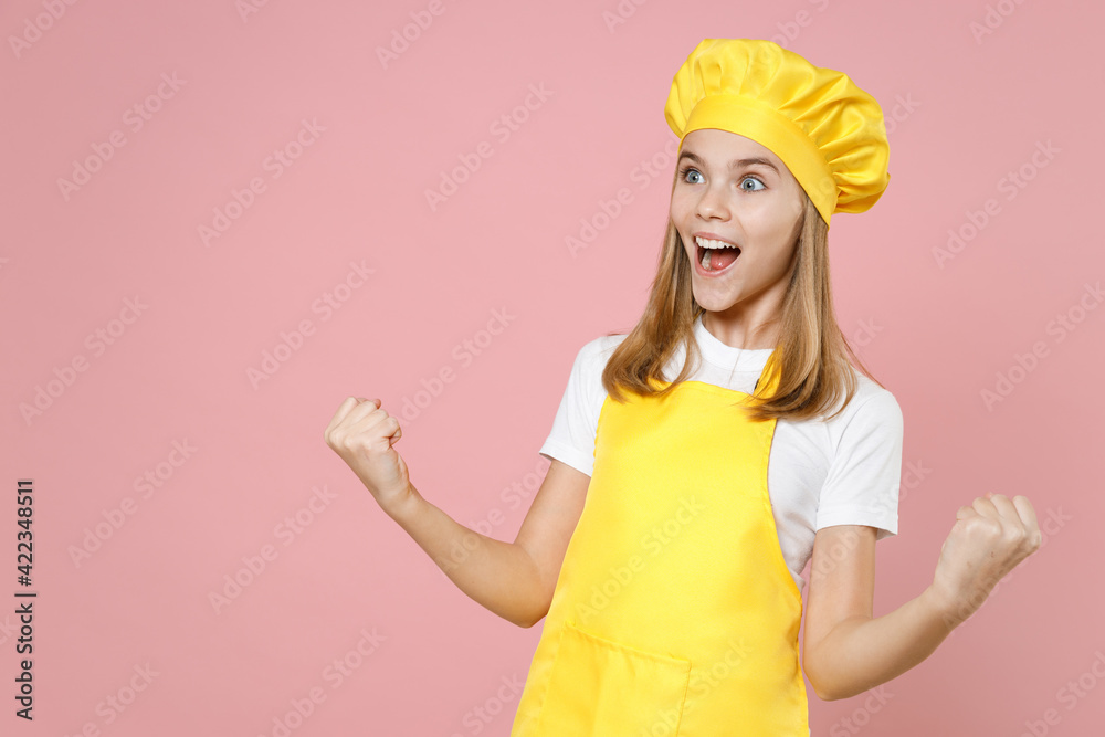 Teen happy girl chef cook confectioner baker housewife mother's helper in yellow apron white t-shirt cap do winner gesture clench fist celebrating isolated on pink background studio Food cake concept. Teen happy girl chef cook confectioner baker housewife mother's helper in yellow apron white t-shirt cap do winner gesture clench fist celebrating isolated on pink background studio Food cake concept.