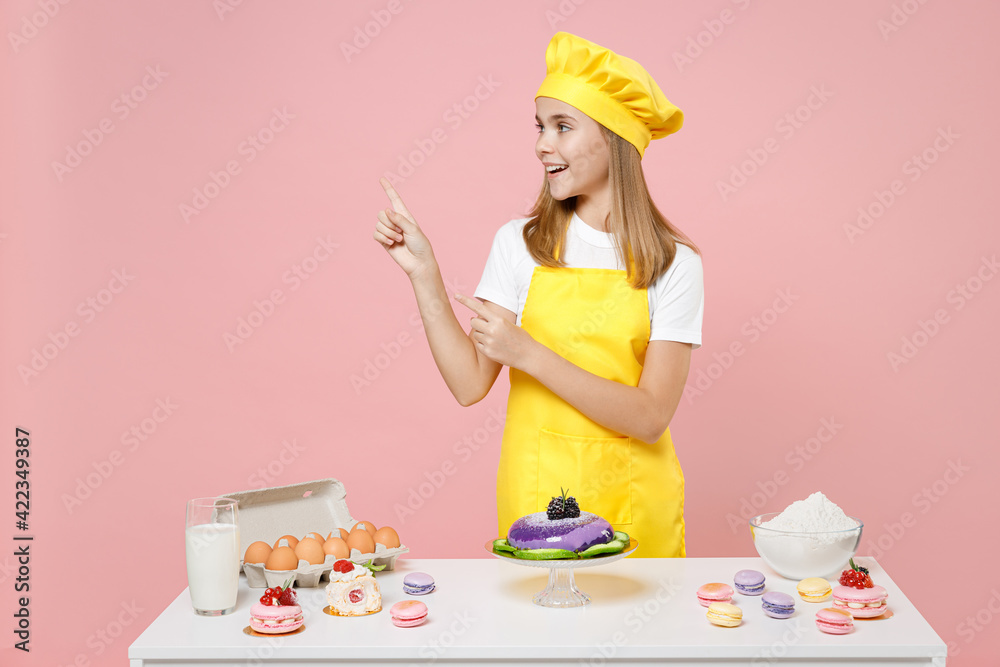 Teen girl chef cook confectioner baker in yellow apron cap at table point index finger aside on workspace mock up isolated on pastel pink background Mousse cake cooking food process workshop concept. Teen girl chef cook confectioner baker in yellow apron cap at table point index finger aside on workspace mock up isolated on pastel pink background Mousse cake cooking food process workshop concept.