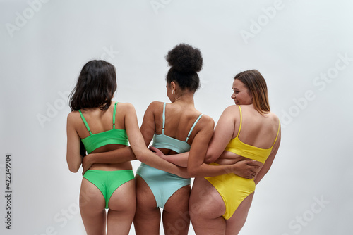 Rear view of three multiethnic women with different body shapes in colorful underwear hugging each other, posing isolated over light background