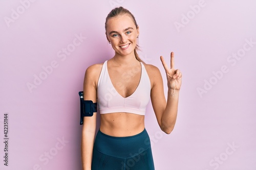 Beautiful blonde woman wearing sportswear and arm band showing and pointing up with fingers number two while smiling confident and happy. © Krakenimages.com