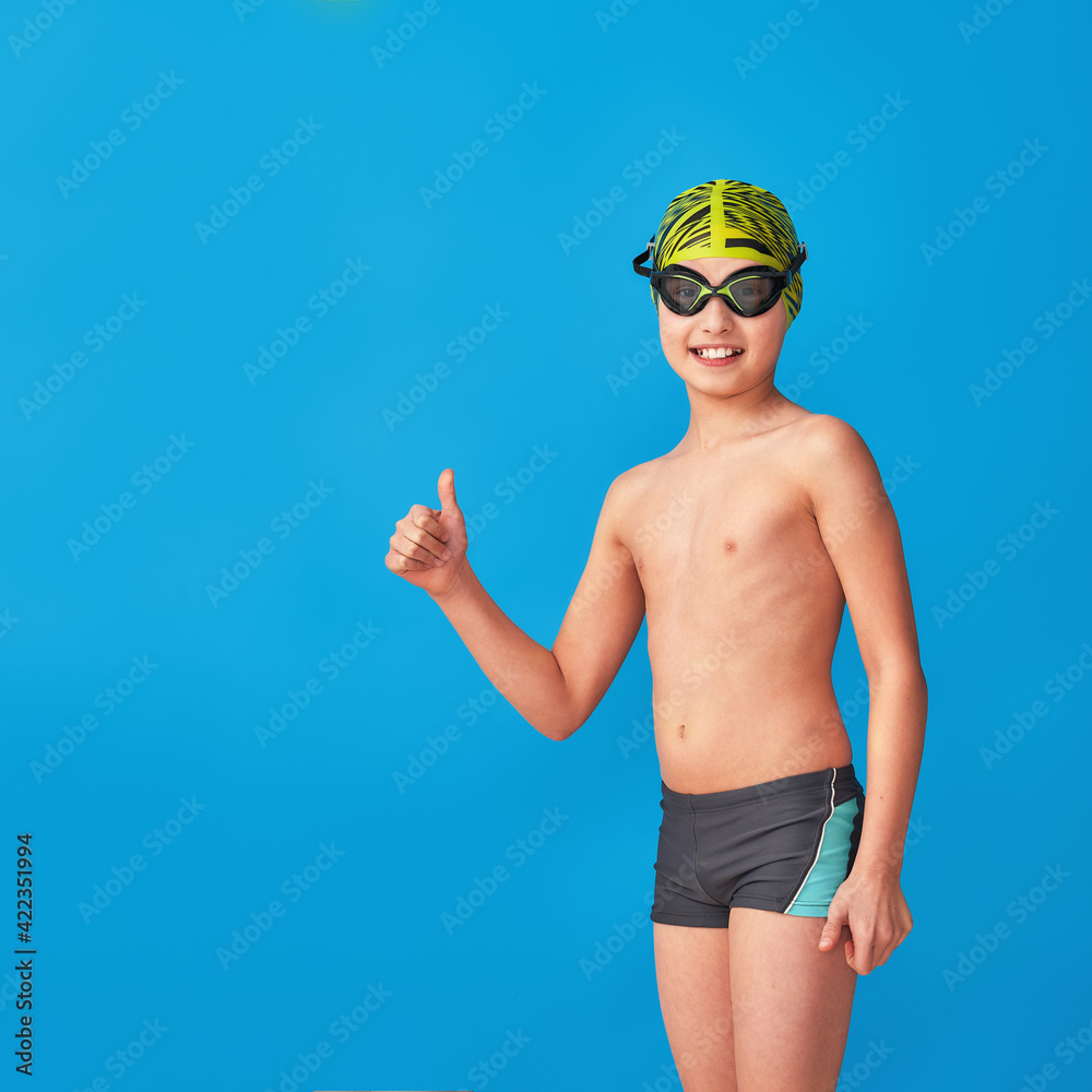 small swimmer in a swimming cap, glasses and a bathing suit on a blue background. the child shows thumbs up, sign of approval, advertising. boy is studying at a sports swimming school. Copy space