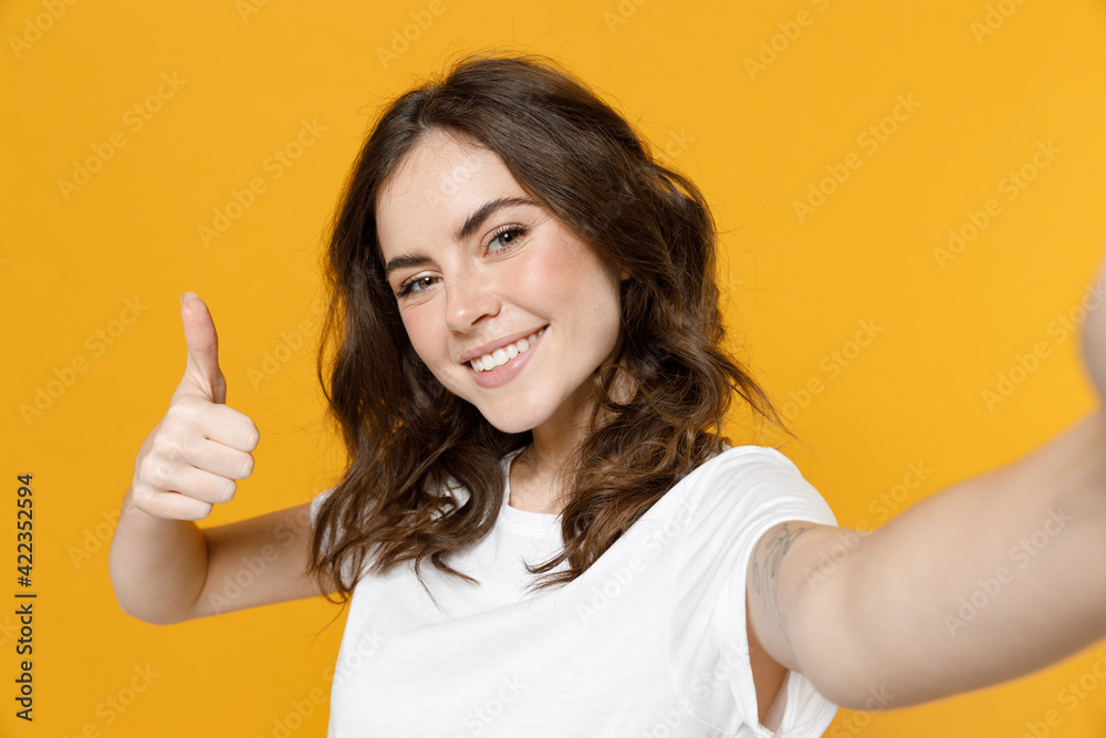 Close up young smiling friendly happy cool caucasian woman 20s in white basic casual t-shirt doing selfie shot on mobile phone show thumb up like gesture isolated on yellow background studio portrait. Close up young smiling friendly happy cool caucasian woman 20s in white basic casual t-shirt doing selfie shot on mobile phone show thumb up like gesture isolated on yellow background studio portrait.