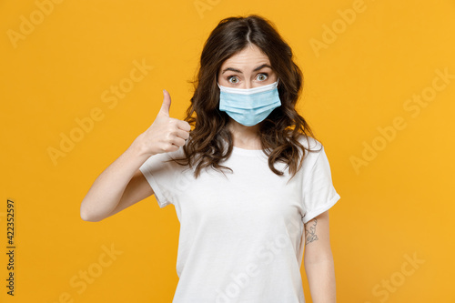 Young woman in white basic blank print design tshirt in sterile face mask to safe from coronavirus virus covid-19 during pandemic quarantine show thumb up gesture isolated on yellow background studio