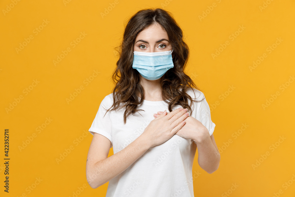 Young kind-hearted good thankful woman 20s wearing white basic t-shirt in sterile face mask from coronavirus virus covid-19 pandemic quarantine put folded hands on heart isolated on yellow background. Young kind-hearted good thankful woman 20s wearing white basic t-shirt in sterile face mask from coronavirus virus covid-19 pandemic quarantine put folded hands on heart isolated on yellow background.