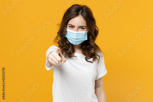 Young woman in white basic blank print design t-shirt in sterile face mask from coronavirus covid-19 pandemic quarantine point index finger camera on you isolated on yellow background studio portrait.