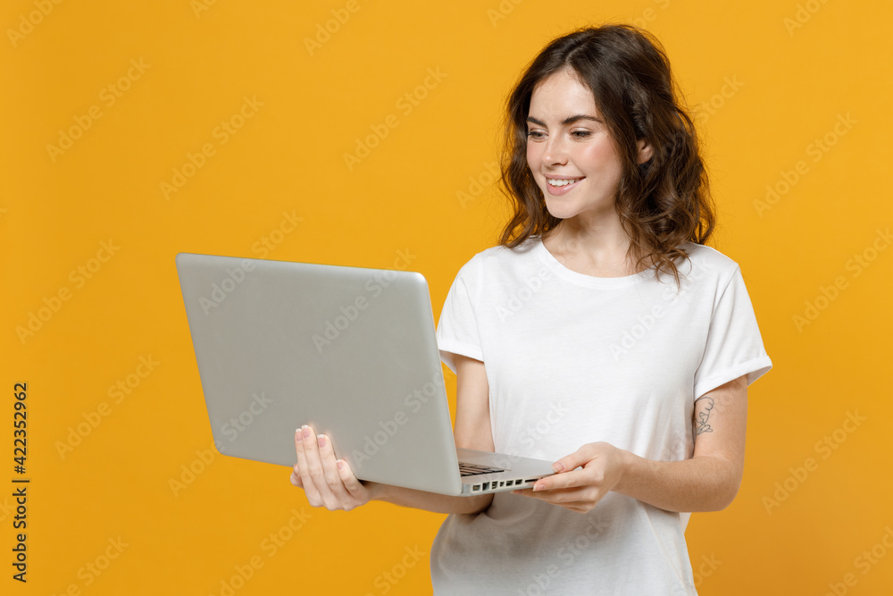 Young smiling happy freelancer copywriter caucasian student woman in white basic t-shirt look camera hold laptop pc computer chat browsing isolated on yellow orange color background studio portrait. Young smiling happy freelancer copywriter caucasian student woman in white basic t-shirt look camera hold laptop pc computer chat browsing isolated on yellow orange color background studio portrait.