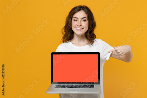 Young smiling freelancer copywriter student woman in white basic t-shirt show point index finger on laptop pc computer with blank screen workspace area isolated on yellow background studio portrait. © ViDi Studio