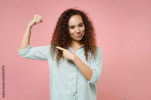 Young black african curly strong sporty fitness woman 20s wear blue shirt point inde finger on biceps muscles on hand demonstrating strength power isolated on pastel pink background studio portrait