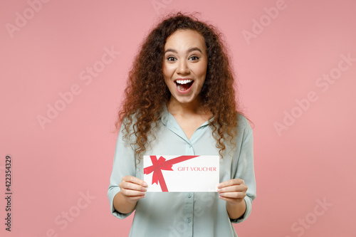 Young black african american surprised shocked cute rich curly woman 20s wearing blue shirt holding gift voucher flyer mock up looking camera isolated on pastel pink color background studio portrait.