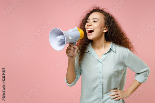 Young black african american surprised excited cute rich curly woman 20s wear blue shirt screaming in megaphone shouting hot news look aside isolated on pastel pink color background studio portrait