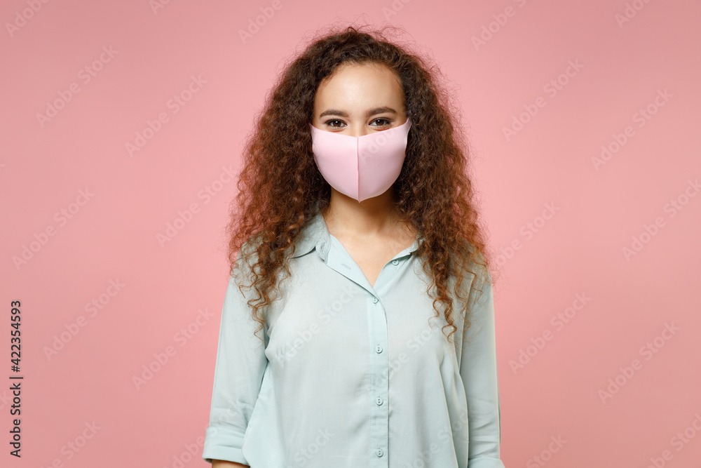 Young black african curly woman 20s wearing blue shirt reusable face mask to safe from coronavirus virus covid-19 during pandemic quarantine isolated on pastel pink color background studio portrait Young black african curly woman 20s wearing blue shirt reusable face mask to safe from coronavirus virus covid-19 during pandemic quarantine isolated on pastel pink color background studio portrait