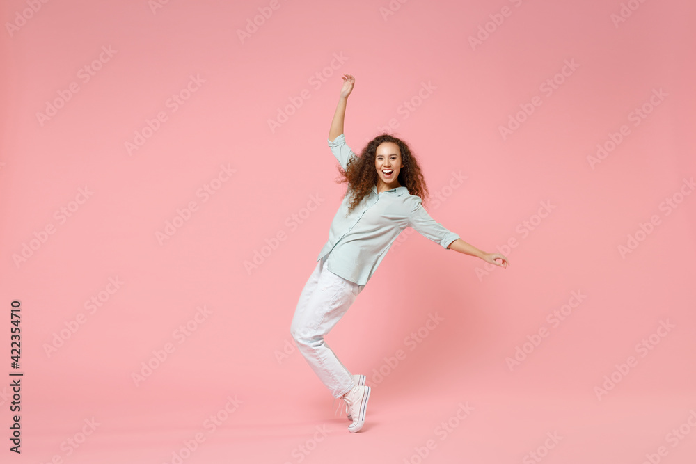 Full length young black african american happy smiling friendly curly woman in blue shirt look camera standing on toes leaning back dancing have fun isolated on pastel pink background studio portrait.