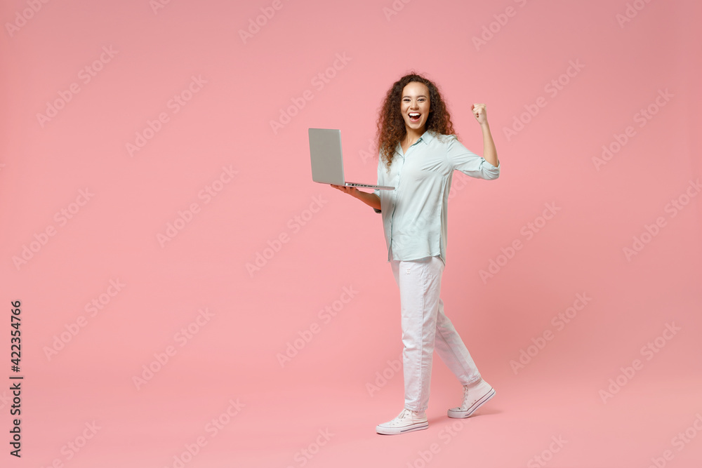 Full length young black african fun happy smiling curly student woman in blue shirt hold laptop pc computer do winner gesture clench fist celebrate isolated on pastel pink background studio portrait Full length young black african fun happy smiling curly student woman in blue shirt hold laptop pc computer do winner gesture clench fist celebrate isolated on pastel pink background studio portrait