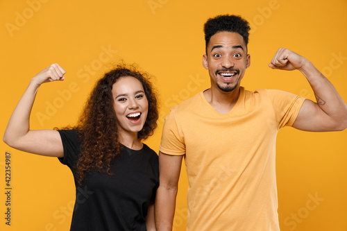 Fényképezés Young couple two american friends strong sporty fitness family african man woman in black t-shirt show biceps muscles on hand demonstrate strength power isolated on yellow background studio portrait