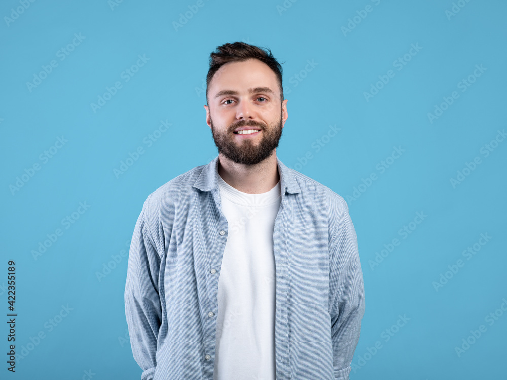 Portrait of cheerful young guy in casual wear posing and smiling at camera on blue studio background