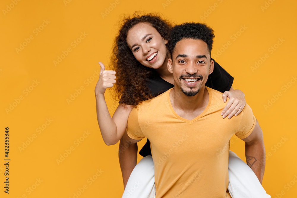 Obraz premium Young couple two together family smiling happy african man woman 20s in black t-shirt giving piggyback ride to joyful sit on back show thumb up gesture isolated on yellow background studio portrait.