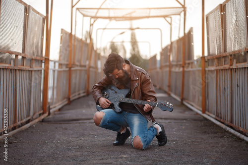A brutal bearded male guitarist in a brown leather jacket and blue jeans sits with his knees on the asphalt and holds a black electric guitar against the background of a road and an iron fence