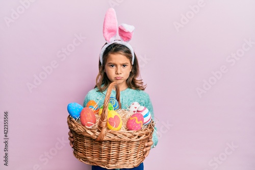 Little beautiful girl wearing cute easter bunny ears holding wicker basket with colored eggs depressed and worry for distress, crying angry and afraid. sad expression.