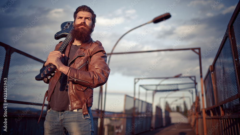 A brutal bearded man with gray hair in a brown leather jacket holds a black electric guitar on his shoulder against the background of an old road A brutal bearded man with gray hair in a brown leather jacket holds a black electric guitar on his shoulder against the background of an old road