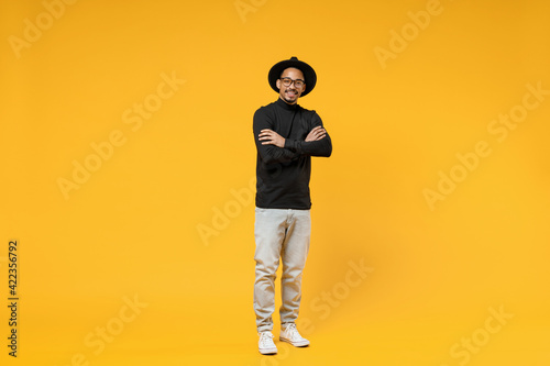 Full length young smiling fashionable fun african american man 20s wearing stylish black hat shirt eyeglasses holding hands crossed folded isolated on yellow orange color background studio portrait