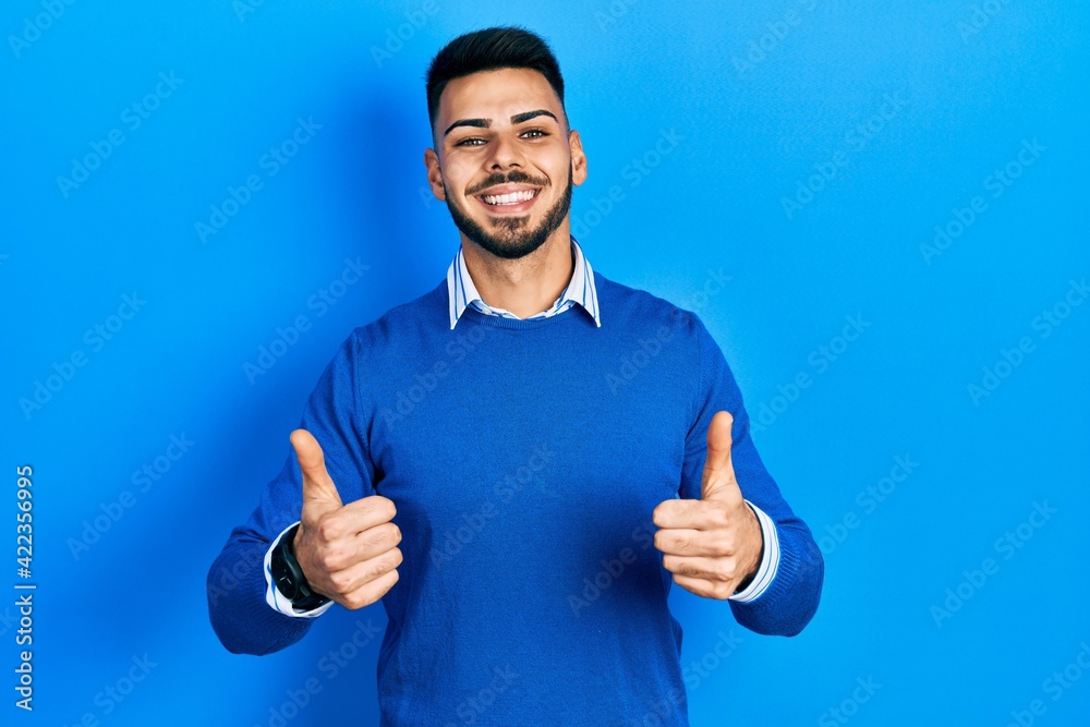 Young hispanic man with beard wearing casual blue sweater success sign doing positive gesture with hand, thumbs up smiling and happy. cheerful expression and winner gesture. Young hispanic man with beard wearing casual blue sweater success sign doing positive gesture with hand, thumbs up smiling and happy. cheerful expression and winner gesture.