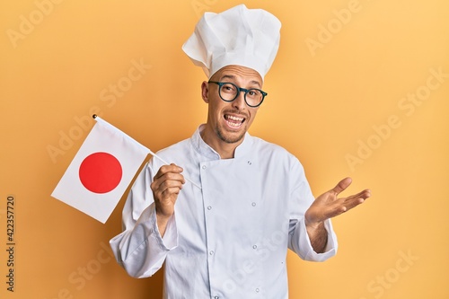 Bald man with beard wearing professional cook uniform holding japan flag celebrating achievement with happy smile and winner expression with raised hand photo