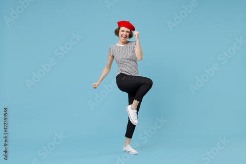 Full length of young overjoyed excited woman with short hairdo in french beret red hat striped t-shirt do winner gesture clench fist raised up leg isolated on pastel blue background studio portrait.