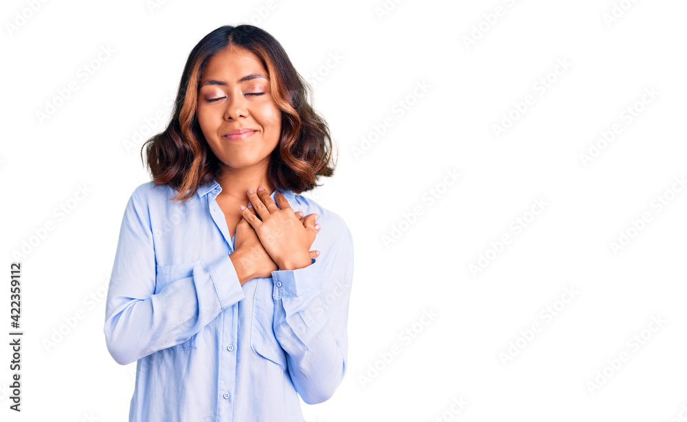 Young beautiful mixed race woman wearing casual business shirt smiling with hands on chest with closed eyes and grateful gesture on face. health concept. Young beautiful mixed race woman wearing casual business shirt smiling with hands on chest with closed eyes and grateful gesture on face. health concept.