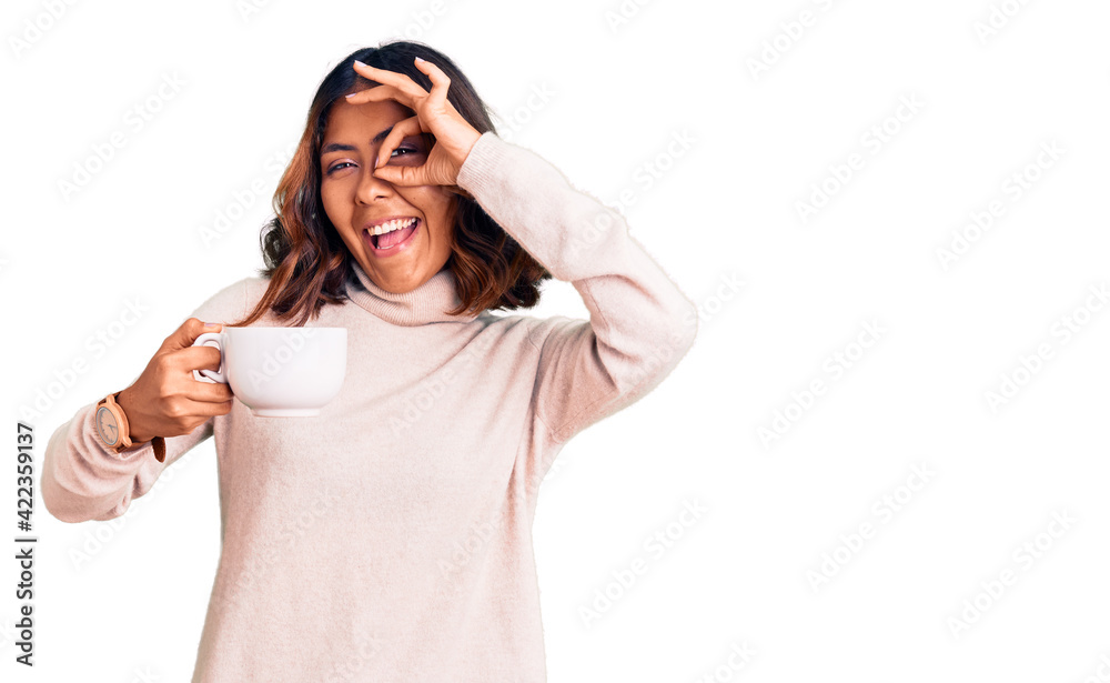 Young beautiful mixed race woman holding a cup of coffee smiling happy doing ok sign with hand on eye looking through fingers Young beautiful mixed race woman holding a cup of coffee smiling happy doing ok sign with hand on eye looking through fingers