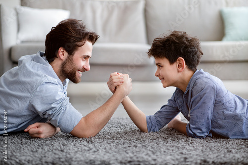 Happy father competing in arm-wrestling with his son, enjoying time together at home, side view photo