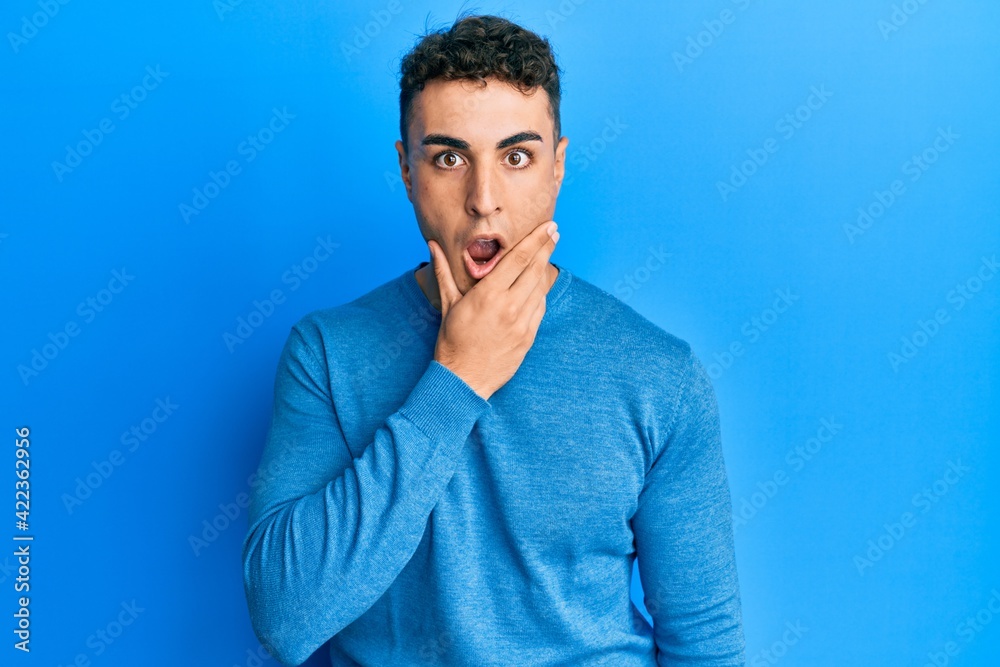 Hispanic young man wearing casual winter sweater looking fascinated with disbelief, surprise and amazed expression with hands on chin Hispanic young man wearing casual winter sweater looking fascinated with disbelief, surprise and amazed expression with hands on chin