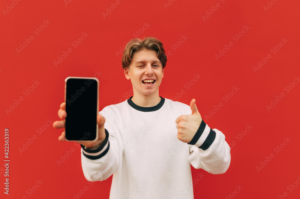 Joyful young man showing thumb up and smartphone screen with camera on red wall background, smiling and looking at camera. Joyful young man showing thumb up and smartphone screen with camera on red wall background, smiling and looking at camera.