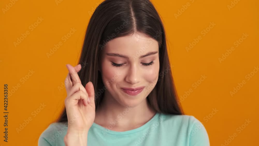 A smiling young woman is crossing her fingers standing isolated over orange background in the studio A smiling young woman is crossing her fingers standing isolated over orange background in the studio