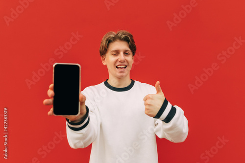Joyful young man showing thumb up and smartphone screen with camera on red wall background, smiling and looking at camera.