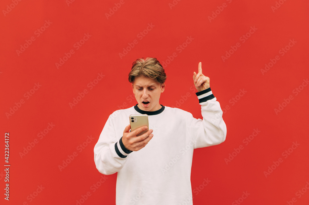 Surprised guy uses a smartphone on a background of a red wall and points his finger up to the place for advertising. Surprised guy uses a smartphone on a background of a red wall and points his finger up to the place for advertising.
