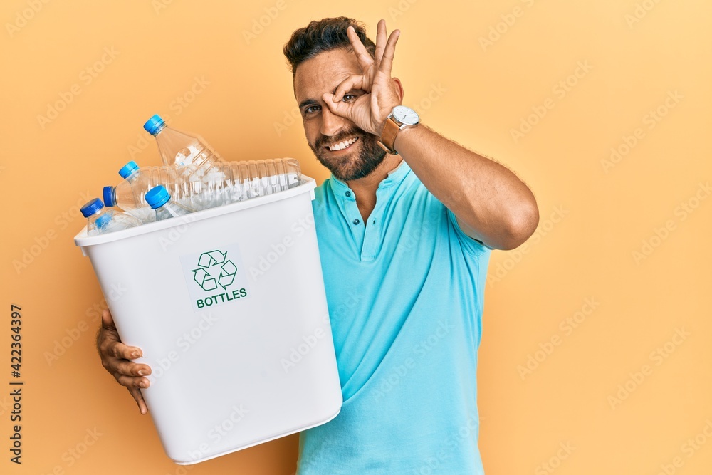 Handsome man with beard holding recycling wastebasket with plastic bottles smiling happy doing ok sign with hand on eye looking through fingers Handsome man with beard holding recycling wastebasket with plastic bottles smiling happy doing ok sign with hand on eye looking through fingers