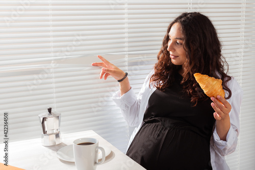 Happy pregnant young beautiful woman eating croissant and looking througt blinds during morning breakfast. Concept of pleasant morning and positive attitude during pregnancy photo