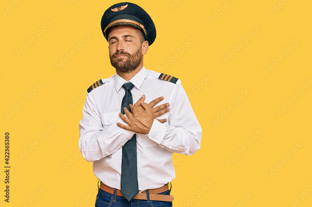 Handsome man with beard wearing airplane pilot uniform smiling with hands on chest with closed eyes and grateful gesture on face. health concept. Handsome man with beard wearing airplane pilot uniform smiling with hands on chest with closed eyes and grateful gesture on face. health concept.