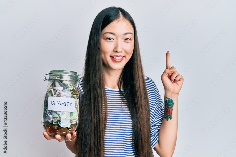 Obraz premium Young chinese woman holding charity jar with money smiling with an idea or question pointing finger with happy face, number one