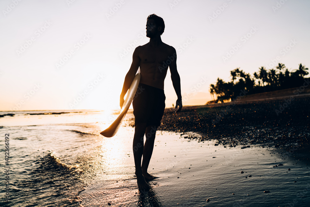 Silhouette of male spending evening for surfing during summer twilight at Carribean sea, amateur man with professional surfboard recreating with sportive activity in Thailand visiting Phuket beach