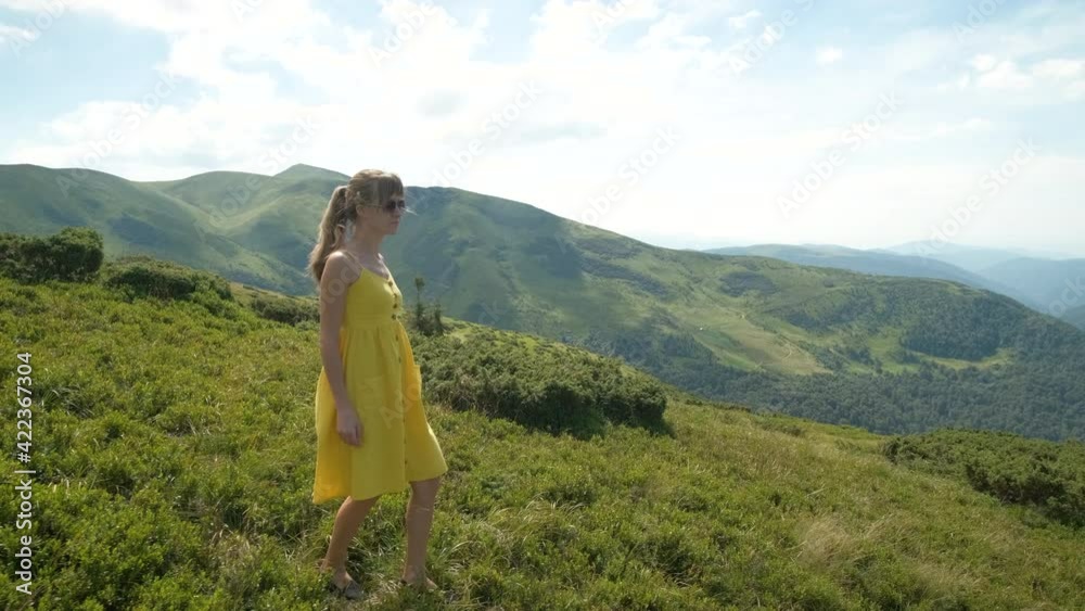 Young happy woman in yellow dress walking on hillside meadow on a windy day in summer mountains. Young happy woman in yellow dress walking on hillside meadow on a windy day in summer mountains.