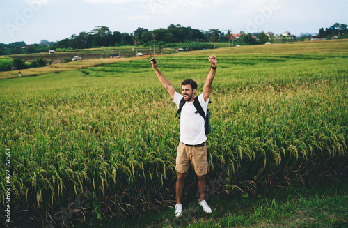 Happy traveler with backpack raising arms in agricultural field