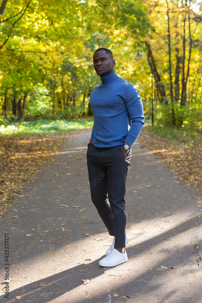 Fototapeta premium Cheerful african american young man in stylish clothes walks in autumn park on sunny warm autumn day. Concept of weekend and outdoor recreation